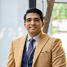 Smiling man in tan blazer and dark tie sitting in a modern office setting with a window background.