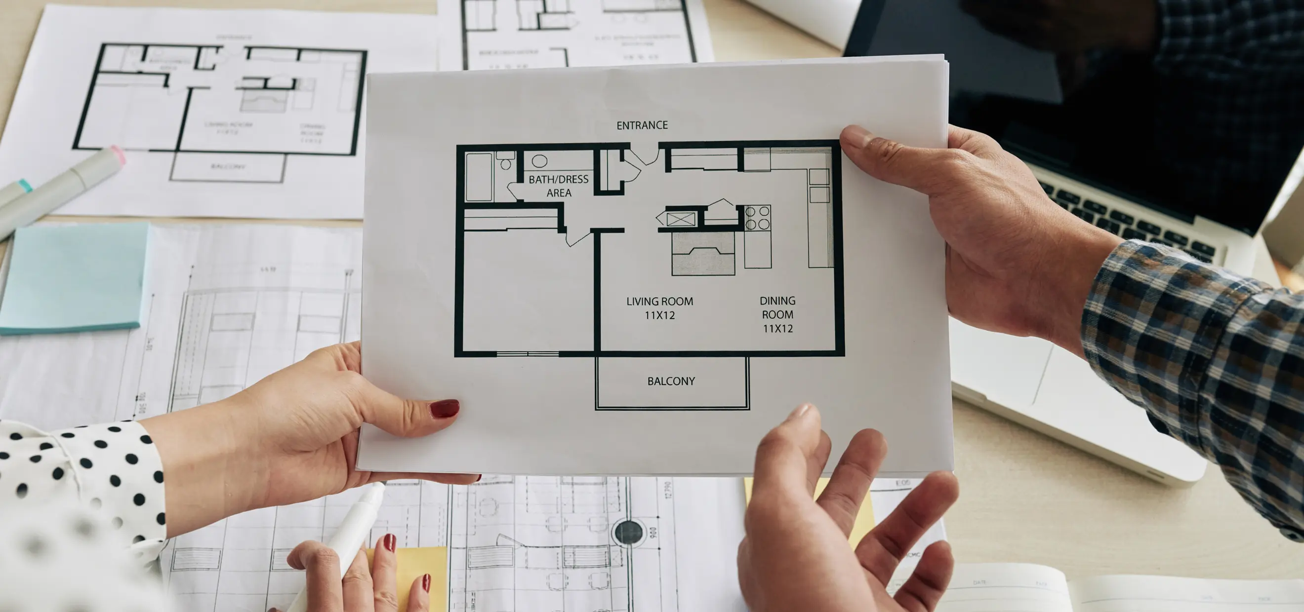 Two people holding a floor plan showing living room, dining room, bath area, entrance, and balcony.