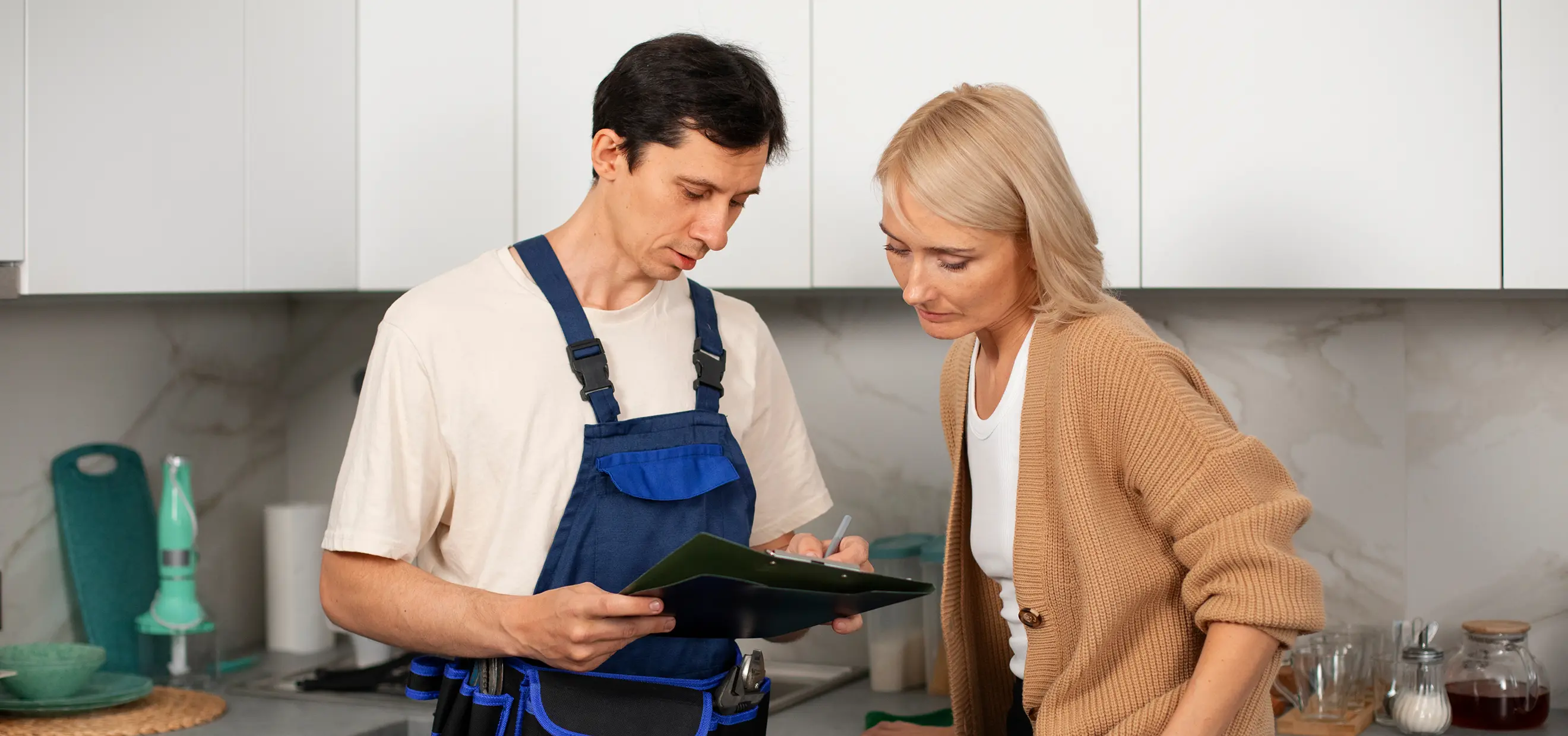 Technician in blue overalls showing clipboard to woman in beige sweater in a kitchen.