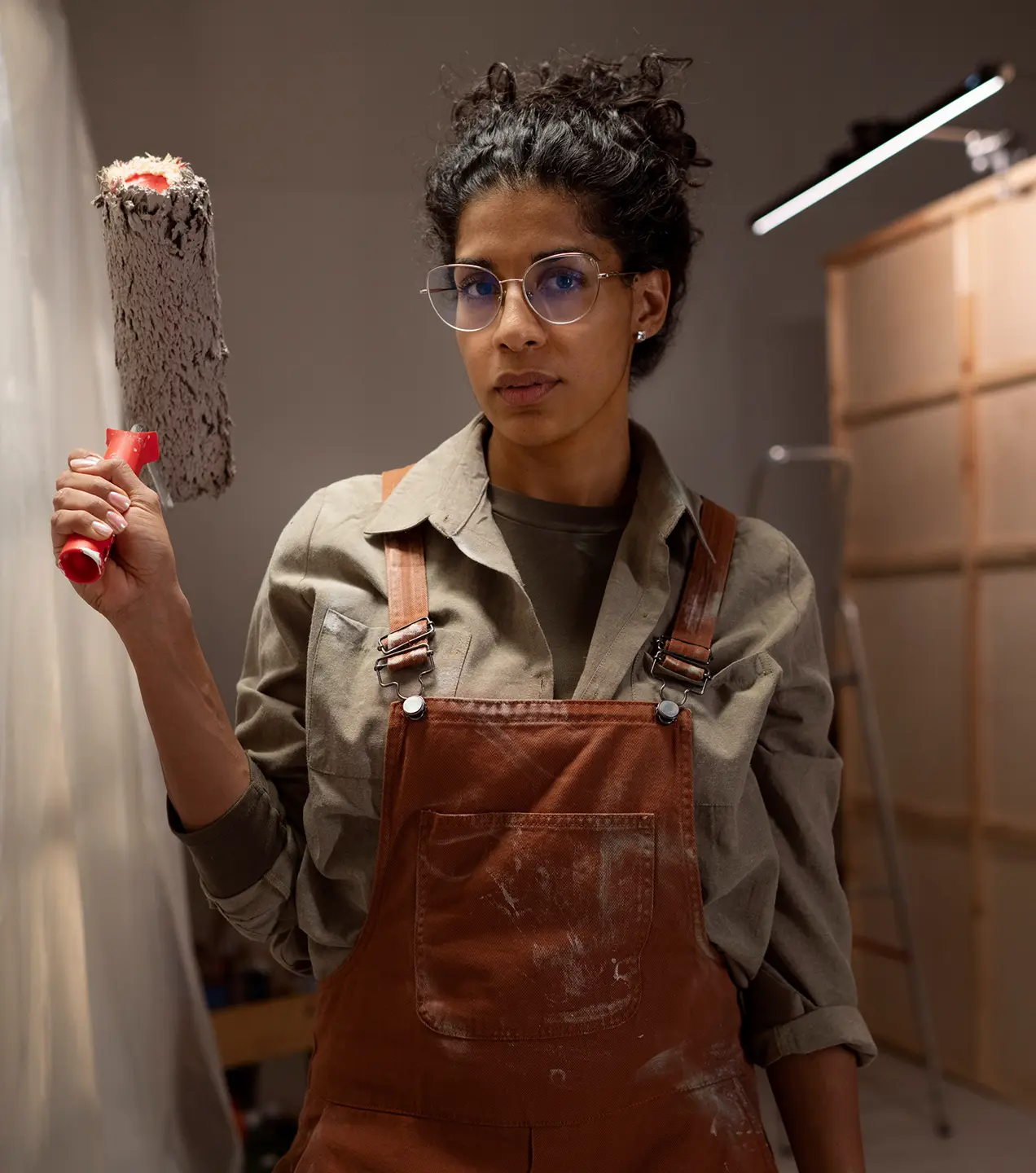 Woman with glasses in paint-stained overalls holding a paint roller indoors.